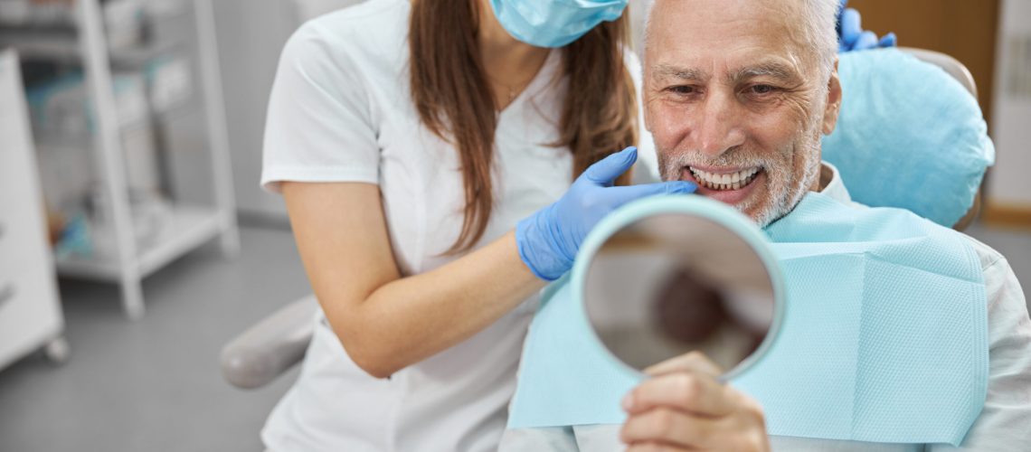 Elderly man and his young female dentist smiling while looking in the mirror in dental clinic