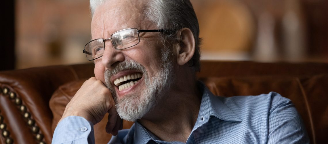 Happy life on pension. Mature aged man retiree wear glasses sit on sofa look aside laugh demonstrate healthy straight teeth prothesis implants. Old man with white smile advertise dental clinic service
