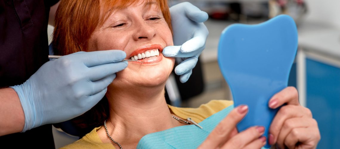 Smiling senior woman with new dental implants sitting in the dental office and looking at the mirror