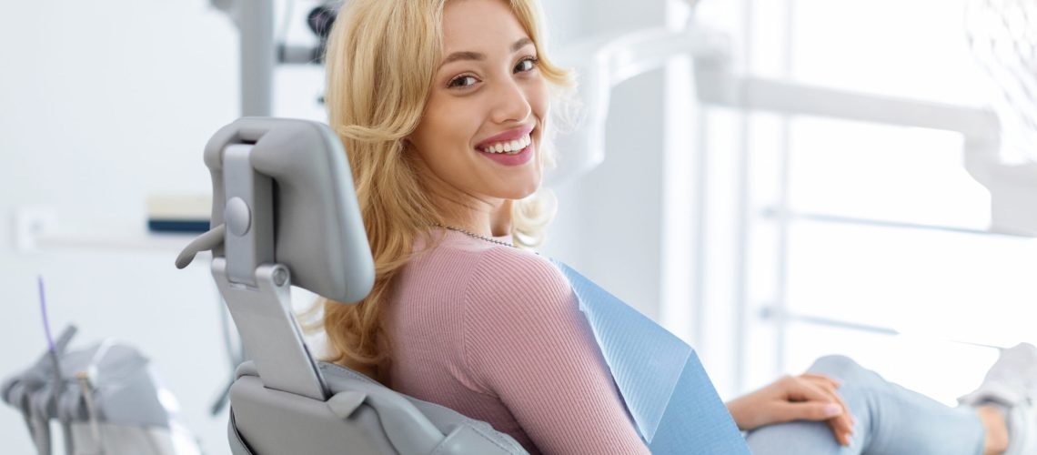 Smiling and relaxed young woman sitting at dental chair in modern clinic, waiting for her dentist. Happy attractive blonde lady attending luxury dental clinic, bacl view, copy space